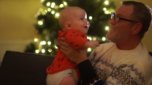 Opa en kleinzoon zitten in de buurt van de kerstboom. De jongen kijkt naar het frame in verrassing. Kerst familie avond — Stockvideo