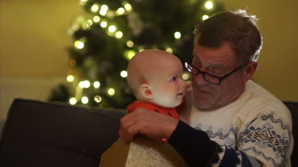 Le retraité ouvre un cadeau de Noël avec son petit-fils. Les lumières brillent hors de la boîte, le bébé a l'air surpris. Premier cadeau — Video