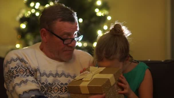 Děda a vnučka u vánočního stromu. Dívka otevírá dar a raduje se. Nová let dárek — Stock video