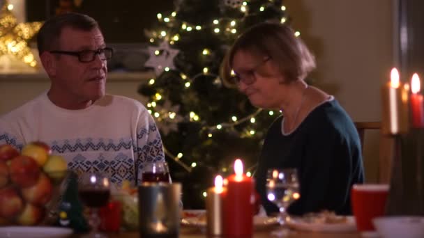 Volwassen man en vrouw spreken aan een tafel met kaarsen tegen de achtergrond van de verlichting van de kerstboom. Eerste datum, bejaarde echtpaar — Stockvideo