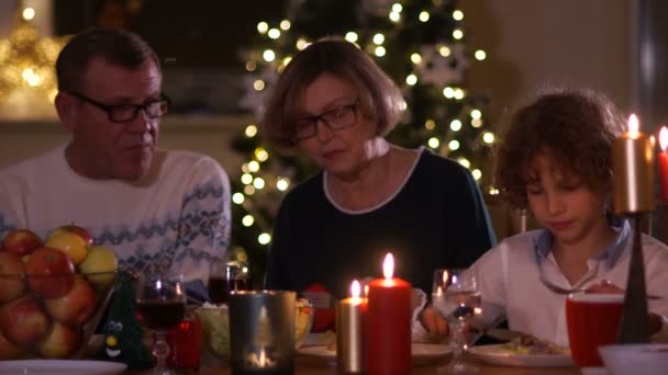 Дедушка и бабушка разговаривают со своим внуком, сидя за праздничным новогодним столом. Ужин при свечах, счастливая семья — стоковое видео