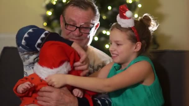 손자와 할아버지 크리스마스에 소파에 앉아 있다. 자매는 산타 모자에 아기를 넣습니다. 놀란된 아기 — 비디오