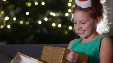 Tatlı bir kız bir Noel hediyesi için bayram yapıyor. Kız öğrenci bir kutu bir hediye ile sarılma yılbaşı ağacı karşısında kanepede oturuyor. Kız dekoratif bir çember ile Noel Baba şapkası takıyor