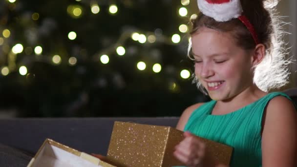 Dulce chica se regocija en un regalo de Navidad. La colegiala está sentada en el sofá frente al árbol de Año Nuevo abrazando una caja con un regalo. La chica lleva un aro decorativo con un sombrero de Santa — Vídeo de stock