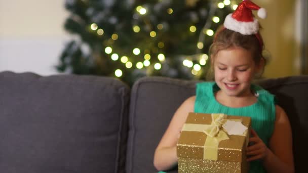 Estudante em um elegante vestido verde e headband com um chapéu de Papai Noel abre um presente. A menina está muito surpreso e encantado, a criança está sentada no sofá perto da árvore de Natal — Vídeo de Stock