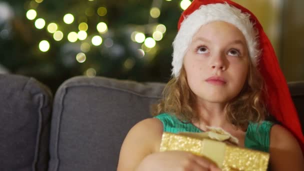 Близкий портрет кареглазая девушка в шляпе Санты с подарочной коробкой в руках. Рождественский праздник, новогодний сюрприз. Девушка мечтательно смотрит вверх — стоковое видео