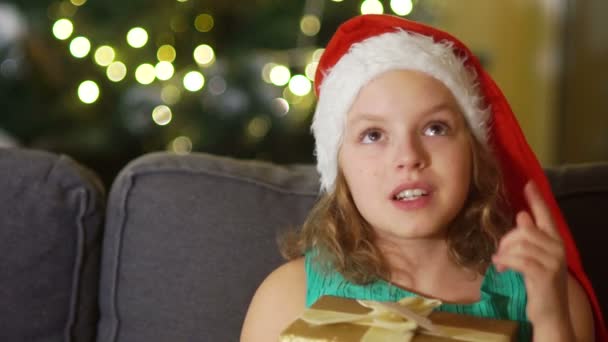 La fille fait un vœu pour Noël. Portrait serré de fille aux yeux bruns en chapeau de Père Noël avec boîte cadeau dans les mains. Fête de Noël, nouvelle surprise de l'année — Video