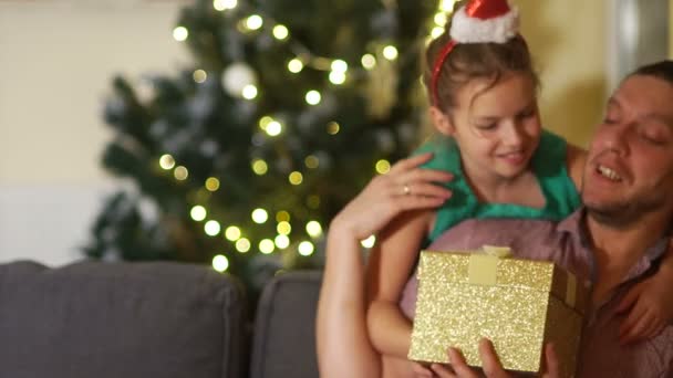 Cute schoolgirl in a rim with a Santa hat gives her father a Christmas gift in a gold box. Happy family christmas — Stock Video