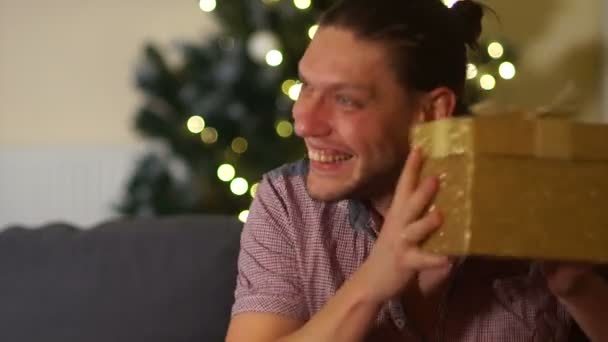 젊은 남자가 크리스마스 선물 크리스마스 트리 근처 압축을 풉니다. 호기심 상자를 떨 려 고 그것을 왜곡, 뚜껑, 유머 — 비디오