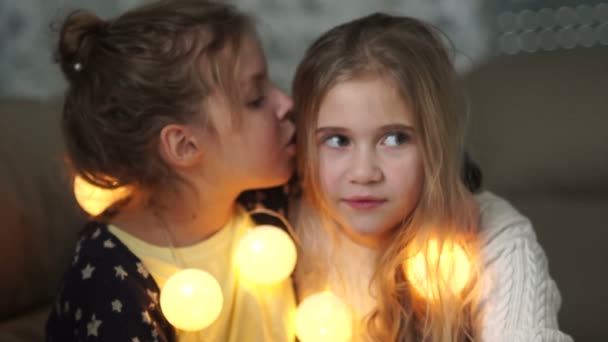Две маленькие подружки делятся секретами. Девушка шепчет сестрам на ухо, оба весело смеются, рождественский вечер, новогодняя гирлянда — стоковое видео