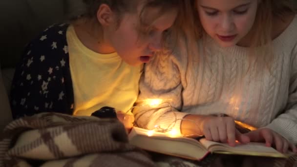 Två flickvänner läsa en bok i sängen på kvällen mot bakgrund av en liten elektrisk krans — Stockvideo