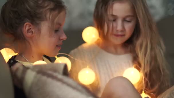 The sisters are covered by a blanket lying in bed with a garland of lights. Girlish friendship — Stock Video