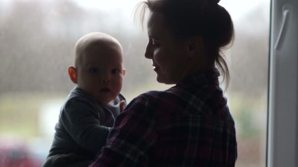 Silhuetten av en ung mor med sin baby. En ung kvinna noggrant håller barnet och ser ut genom fönstret. Väntar på pappa — Stockvideo