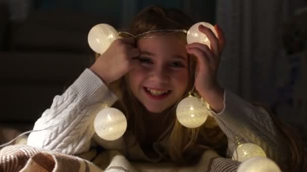Porträt eines netten Mädchens mit Weihnachtsbeleuchtung. Kind liegt auf dem Boden und spielt mit Weihnachtsgirlanden, kreatives Dekor — Stockvideo