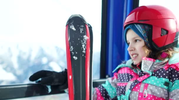 Nahes Porträt vor dem Hintergrund verschneiter Berge. Teenager mit Helm und Skiern erklimmt einen Berg in der Skiliftkabine. Skigebiet, Familienskifahren — Stockvideo