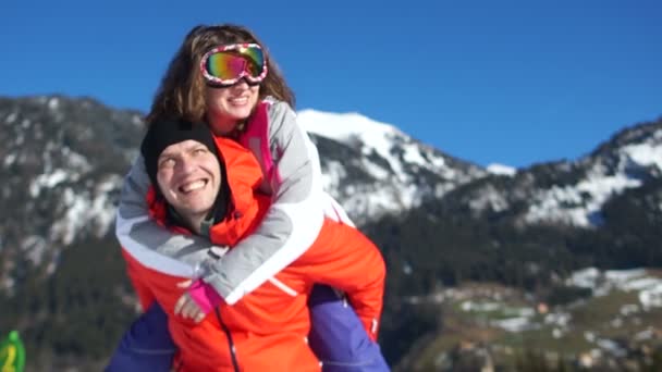 在山上滑雪度假的幸福夫妇。穿着滑雪夹克和护目镜的男人和女人。那个女孩在他的背上跳了个男人 — 图库视频影像