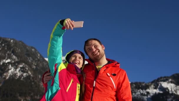 Couple de skieurs en vestes de ski rouges faisant selfy avec smartphone contre le ciel bleu et les montagnes d'hiver. Vacances dans la station de ski — Video
