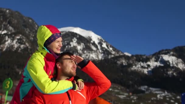 Casal encantador está descansando nas montanhas no inverno. Um homem e uma mulher de casacos de esqui vermelhos no fundo de encostas de montanha nevadas — Vídeo de Stock