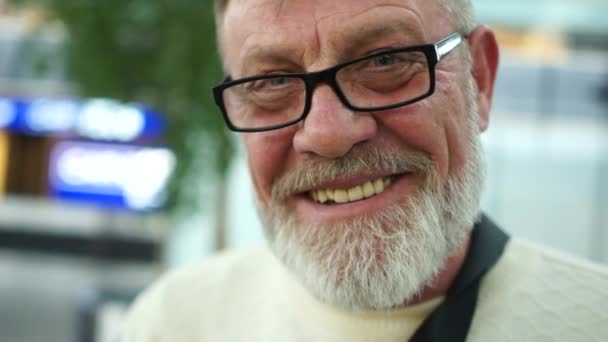 Retrato cercano de un hombre maduro con gafas y barba gris. Pensionista sonriendo sinceramente en el aeropuerto, centro comercial, mirando a la cámara — Vídeos de Stock