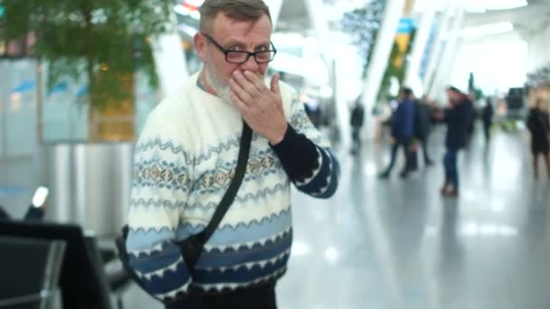 Hombre maduro con una barba gris en un suéter de punto y gafas en el aeropuerto o estación de tren. El pensionista se despide y se va llevando una maleta sobre ruedas detrás de él — Vídeo de stock