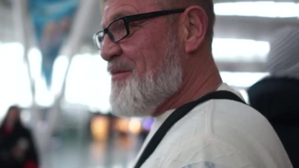 Porträt eines graubärtigen Touristenführers, der sich am Flughafen räkelt. der Reisende trägt eine Brille und einen weißen Pullover — Stockvideo