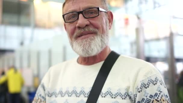 Laughing elderly man with glasses and a gray beard at the airport. Close portrait, waiting for the flight — Stock Video