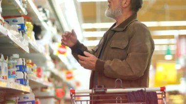 Yaşlı bir adam bir süpermarkette peynir süt bölümünde seçmek çalışıyor. Gülümsüyor ve belgili tanımlık smartphone görünüyor. Sağlıklı gıda