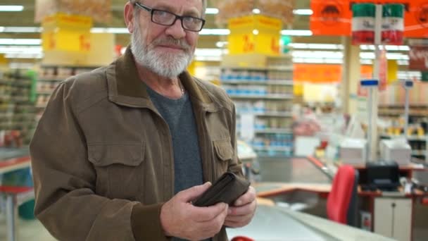 El hombre con barba gris y gafas sostiene una cartera entera en un supermercado y mira a la cámara sonriendo. Retrato cerrado — Vídeos de Stock