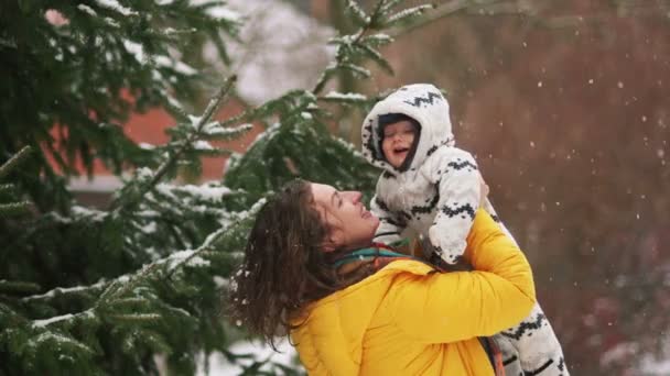 Bambino di otto mesi in passeggiata con sua madre. Donna raddrizza il suo berretto bambino. Sta nevicando, sullo sfondo di abete rosso e parete — Video Stock