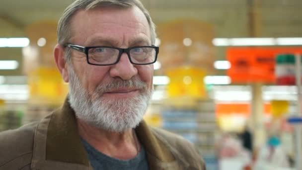 Reifer Rentner im Supermarkt, nahes Porträt, trauriges Lächeln. der Mann hat einen gepflegten grauen Bart, blaue Augen und eine optische Brille, Falten sind sichtbar — Stockvideo