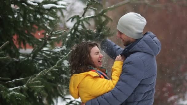 Lykkelig ungt par under gran som morer seg i vinterparken. En mann rister snø av en gren av en kvinne. Elskere spøker – stockvideo