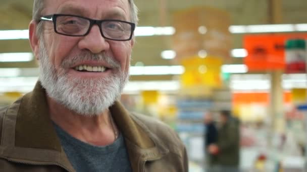 El hombre en un supermercado sonríe, agita su mano con una cartera de molestias. Barba gris y gafas, las arrugas son visibles. Concepto de pobreza — Vídeos de Stock