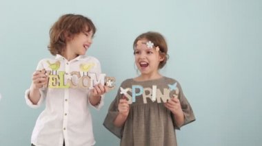 Bahar kavramı. Stüdyo portre sevimli okul çocukları ile ahşap işaretler. Bahar tatili, pastel renkler. Oğlan beyaz bir gömlek ve kız bir bej elbise ve çiçekli bir çelenk giyiyor