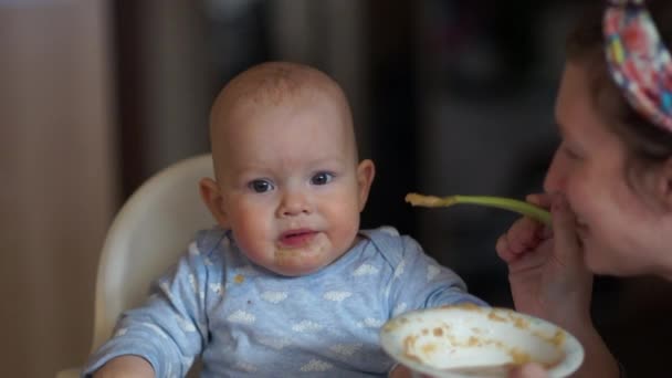 El primer bebé que alimenta. El niño se sienta en una silla alta, la madre le da una cucharada de puré de frutas. El niño no quiere comer, ensuciado en la comida — Vídeos de Stock