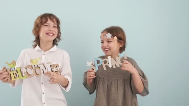 Ένα αγόρι και ένα κορίτσι, άλμα, χορό και να διασκεδάσουν στο studio σε μπλε φόντο με άνοιξη διακόσμηση στα χέρια τους. Ένα αγόρι σε ένα λευκό πουκάμισο και ένα κορίτσι σε ένα μπεζ φόρεμα με ένα στεφάνι στο κεφάλι του — Αρχείο Βίντεο