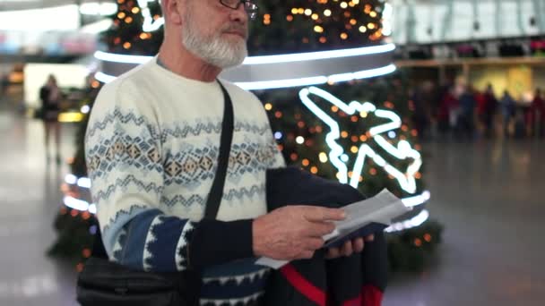 Ein Mann mit grauem Bart wartet an Weihnachten und im neuen Jahr auf seinen Flug zum internationalen Flughafen. Er hält seinen Pass und die Fahrkarten in der Hand und schaut auf den Fahrplan — Stockvideo