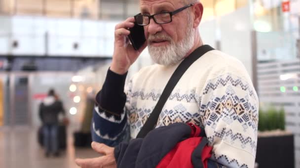 Дід з мобільним телефоном в руці розмовляє в аеропорту. Чоловік строгий і серйозний — стокове відео