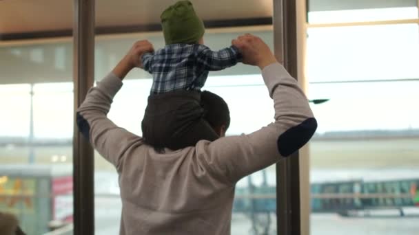 Мужчина держит своего годовалого ребенка на плечах. Отец и сын смотрят на самолет через окно в аэропорту. счастливая семья, отец-одиночка, день отца — стоковое видео