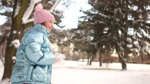 Девушка в синей куртке и розовой шляпе бросает снежок в своего отца. Зимний солнечный день в парке, счастливые семейные праздники — стоковое видео