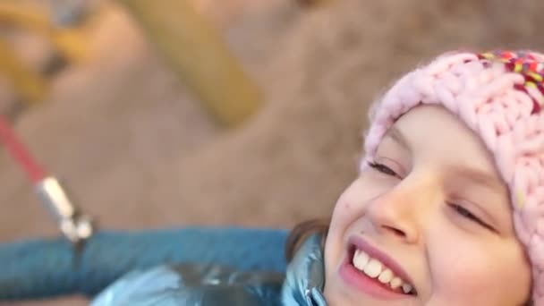 Schulmädchen schaukeln in den Winterferien auf einer Schaukel auf dem Spielplatz. Das Mädchen trägt eine rosa Strickmütze. Porträt aus nächster Nähe — Stockvideo