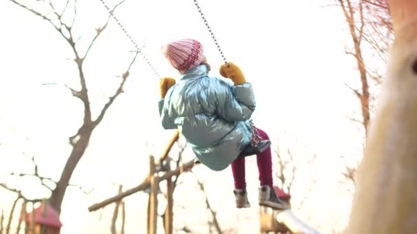 Милая девочка-подросток, качающаяся на качелях на детской площадке во время зимних каникул. Девушка носит розовую вязаную шляпу — стоковое видео
