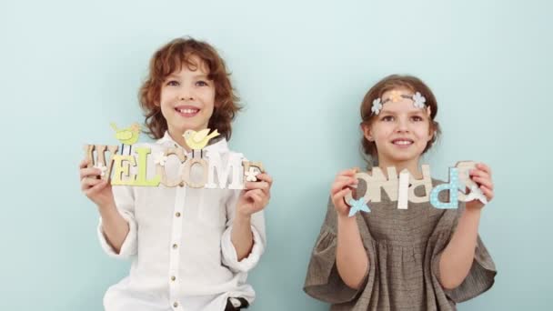 Retrato de estudio de niños de escuela lindos con carteles de madera. Vacaciones de primavera, colores pastel. El chico lleva una camisa blanca, y un vestido beige y una corona de flores. — Vídeos de Stock