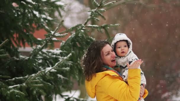 Mulher nova encantadora em uma jaqueta amarela anda com o bebê no macacão. Está nevando, a mãe mostra seu filho, a criança parece surpreso — Vídeo de Stock