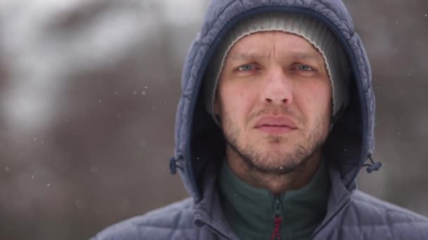 Close portrait of a stern man with a beard in winter clothes and with a hood on his head in a snowstorm against the background of a winter forest. Copy space left — Stock Video