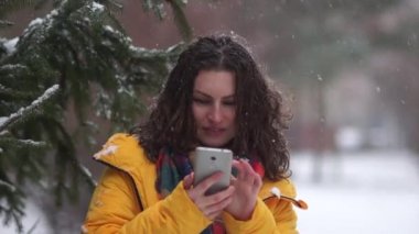 Kar yağışı sırasında bir akıllı telefon ile parkta yürüyen bir kıvırcık esmer portresi kapatın. Karla kaplı ladin, bir kız bir sarı ceket aşağı zemine karşı