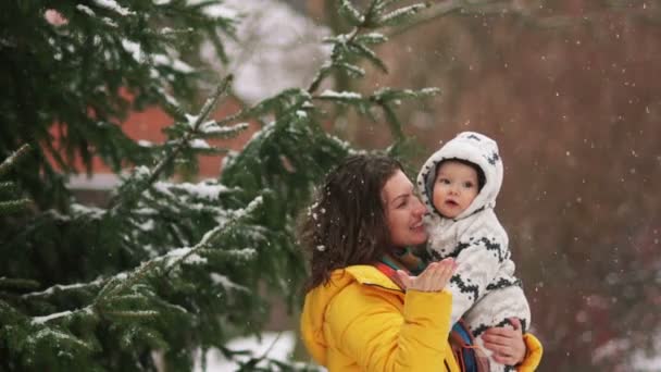 Madre e hijo admiran la primera nieve en el parque cerca del abeto. La niña está vestida con una chaqueta amarilla, el niño lleva un mono. Día de las Madres — Vídeos de Stock