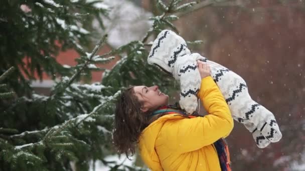 Gelukkig moeder op een winter lopen in het park met de baby. Het sneeuwt, de vrouw verhoogt het kind tot lachen en hem kussen. Het kind is gekleed in een warme jumpsuit, het meisje draagt een gele jas — Stockvideo