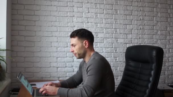Stilig affärsman arbetar med laptop i office. En man sitter i en skinnfåtölj mot en vit tegelvägg — Stockvideo