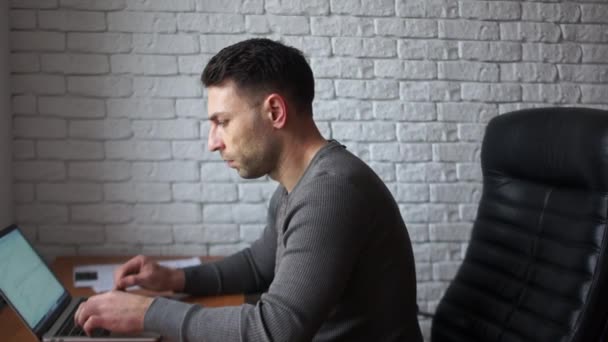 Un giovane programmatore stanco in ufficio sta lavorando con un computer portatile. Un uomo siede su una sedia di pelle contro un muro di mattoni bianchi — Video Stock