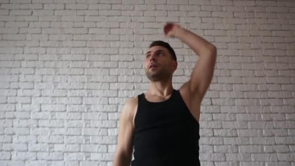 Fitness man in een zwart t-shirt aan het warmrijden in gym. Atletische brunette jongeman met een baard doet oefeningen op de achtergrond van een witte bakstenen muur — Stockvideo